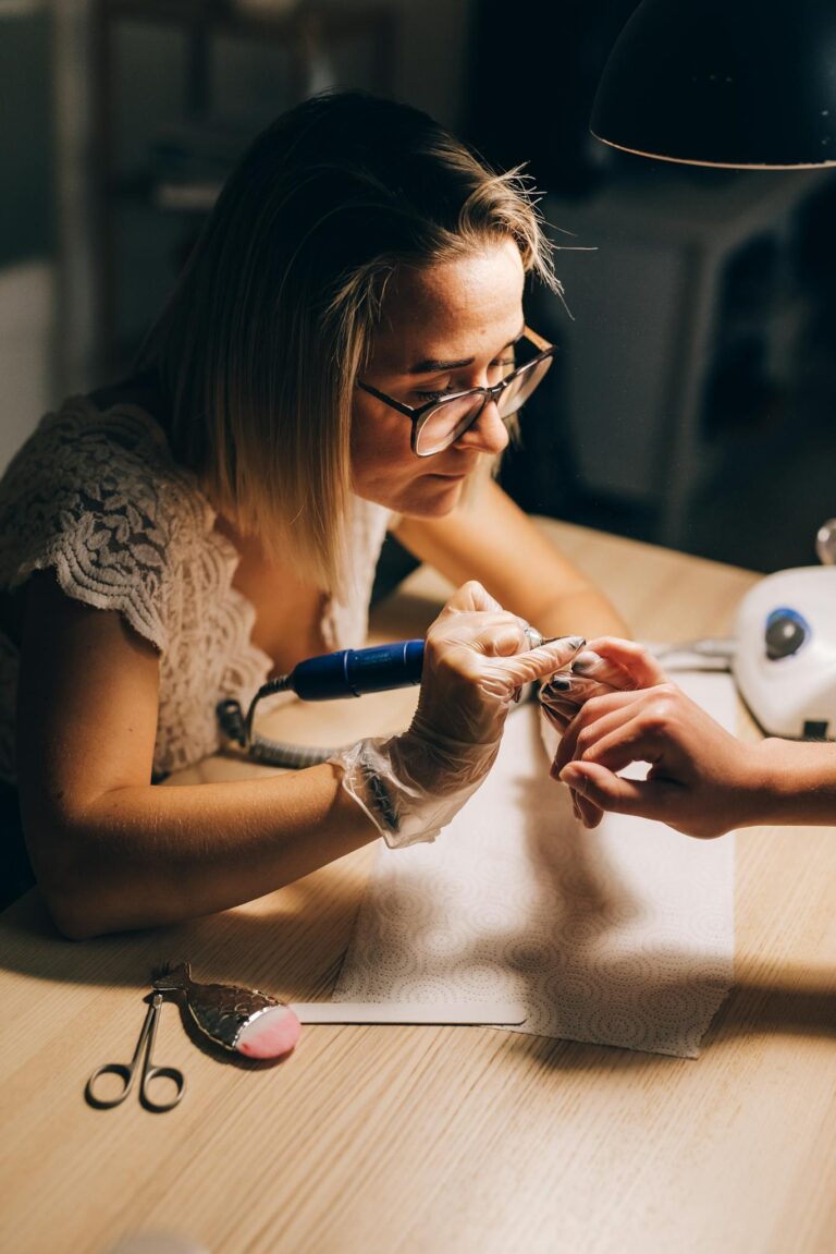 High angle of concentrated female master in glasses and gloves manicuring hand with drill of faceless client