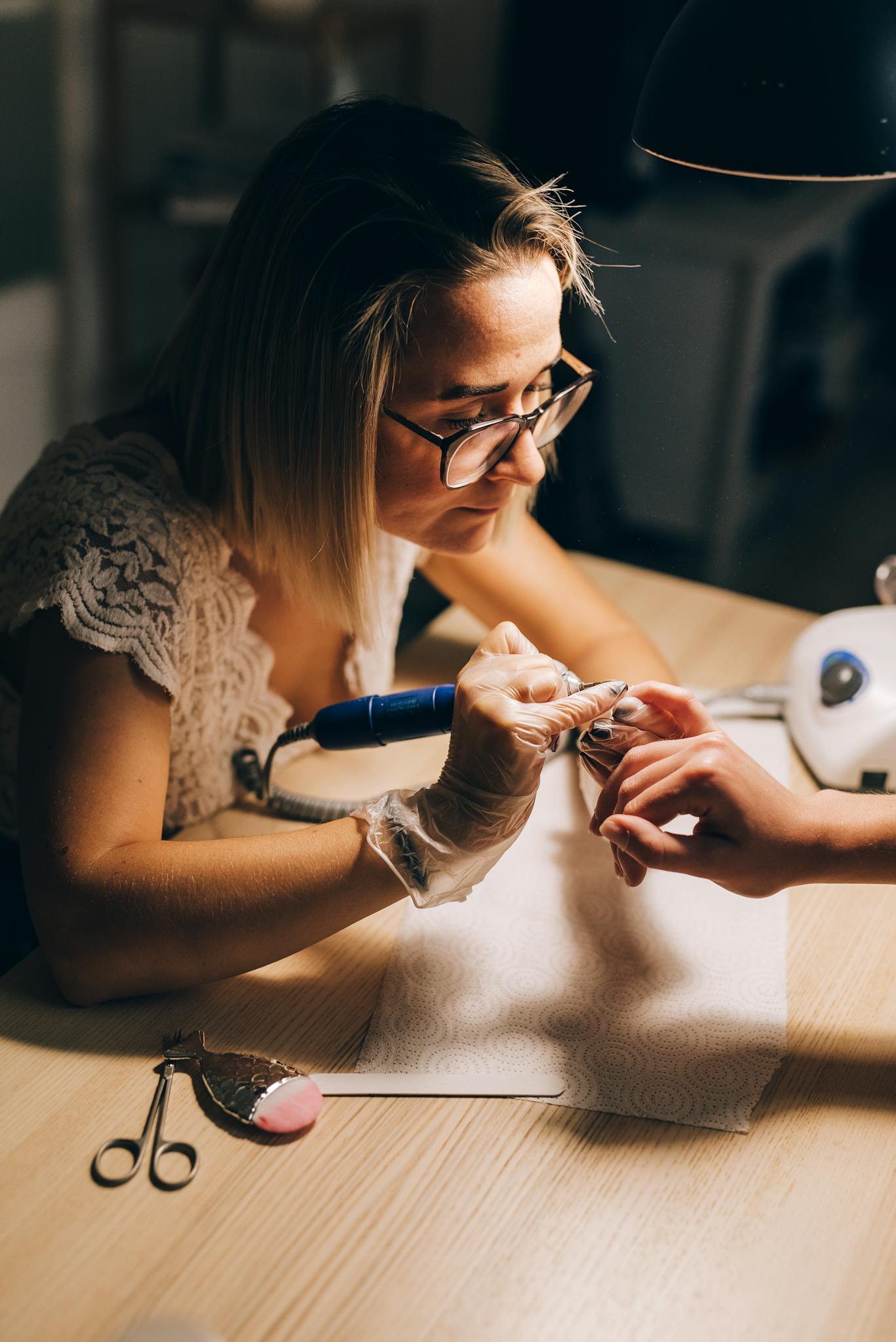 High angle of concentrated female master in glasses and gloves manicuring hand with drill of faceless client